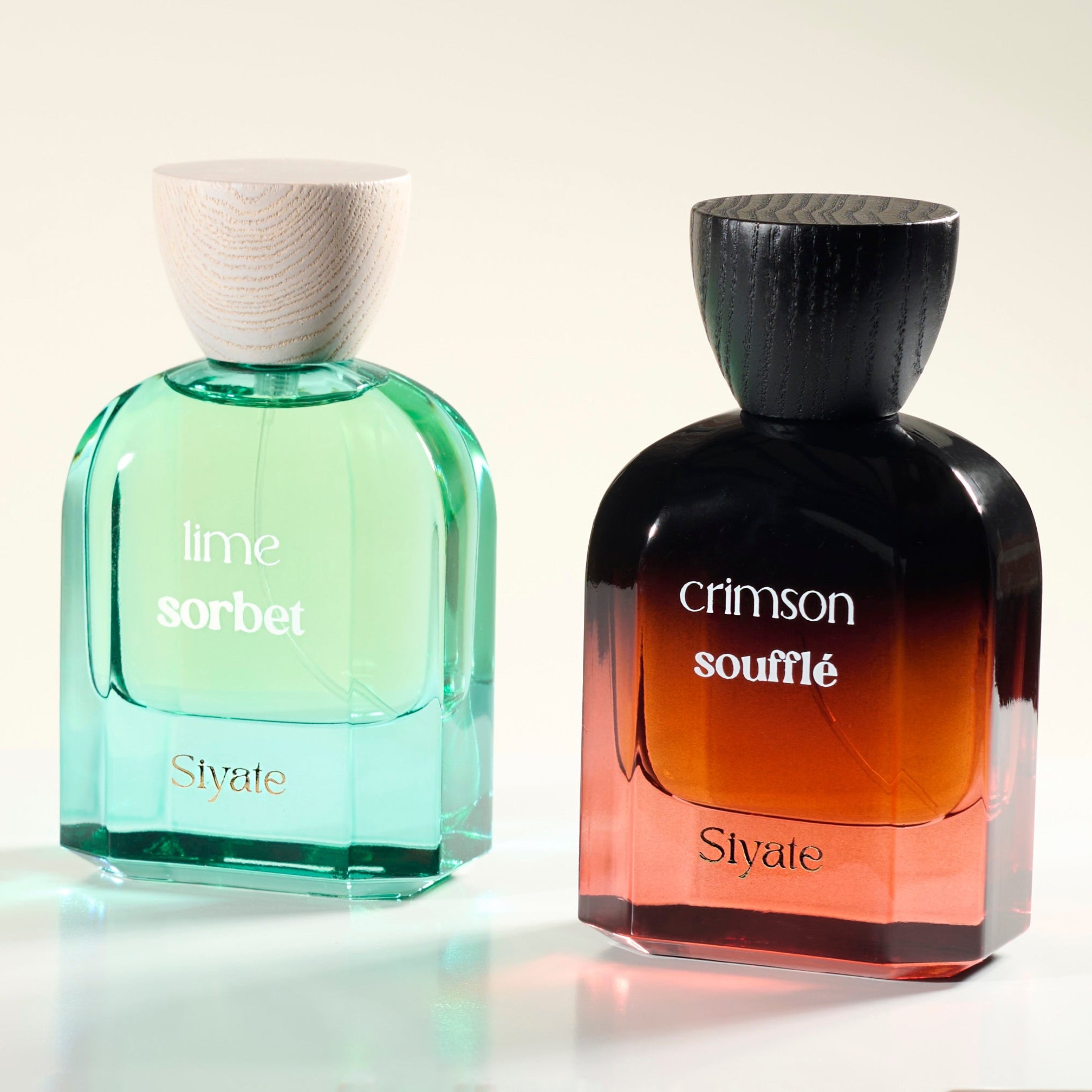 Two bottles of Stvate perfume on a white reflective surface against a light beige background. The left bottle is lime sorbet with a light green translucent glass body and a white cap.