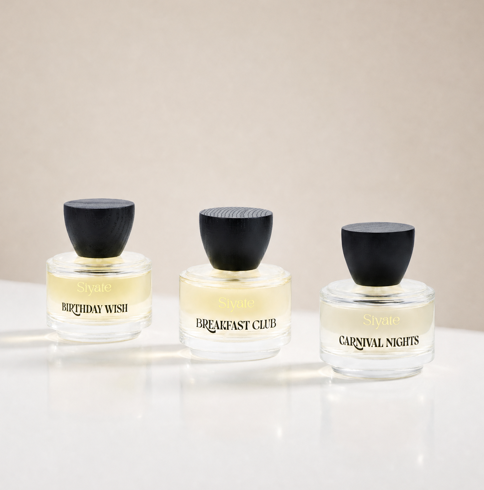Three small clear glass perfume bottles with black caps arranged on a white surface against a warm beige background. The bottles are labeled 'Sydne' and show fragrance names 'BIRTHDAY WISH', 'BREAKFAST CLUB', and 'CARNIVAL NIGHTS'.