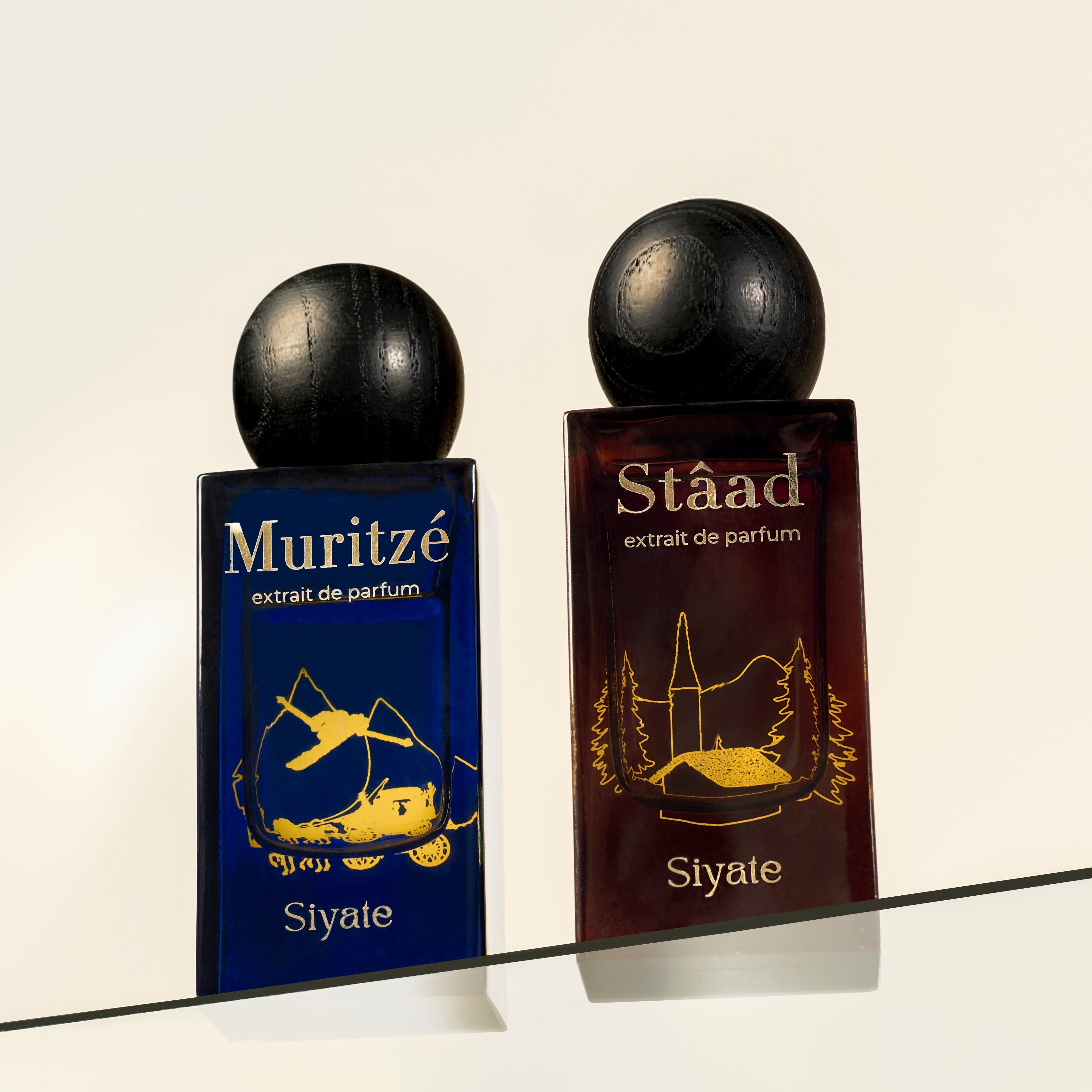 Two rectangular perfume bottles with spherical black caps displayed on a glass surface against a light beige background. The left bottle is dark blue with gold artwork and reads 'Muritze extrait de parfum Siyate.