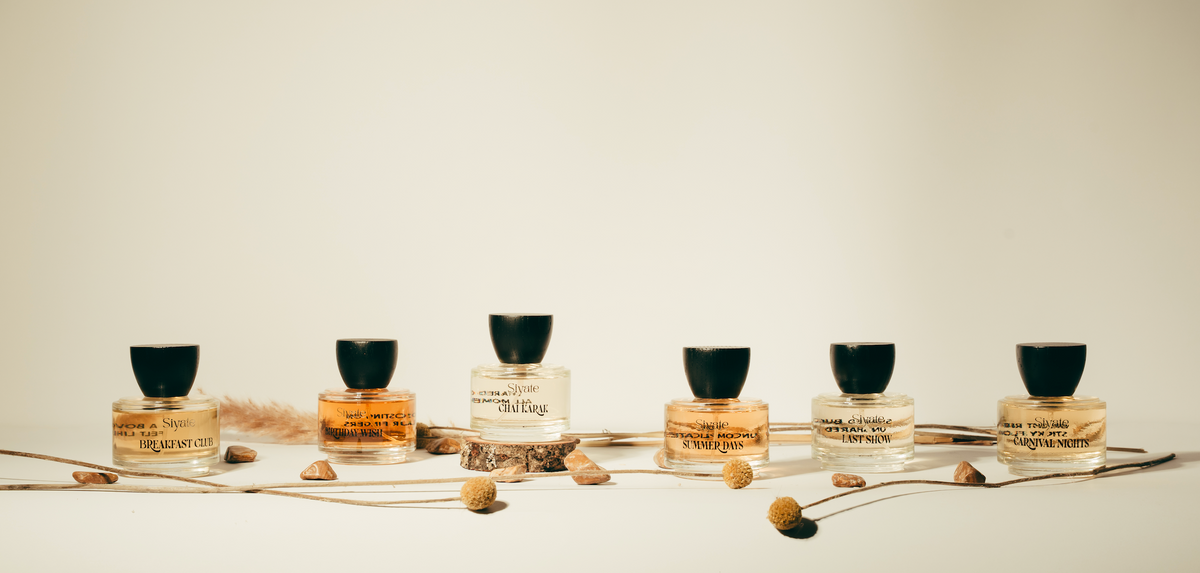 Six small glass perfume bottles with black caps are lined up on a light beige background, decorated with dried flowers and stems. The bottles contain clear liquid and display labels including 'Scent of the Wild' and 'The Fog'.