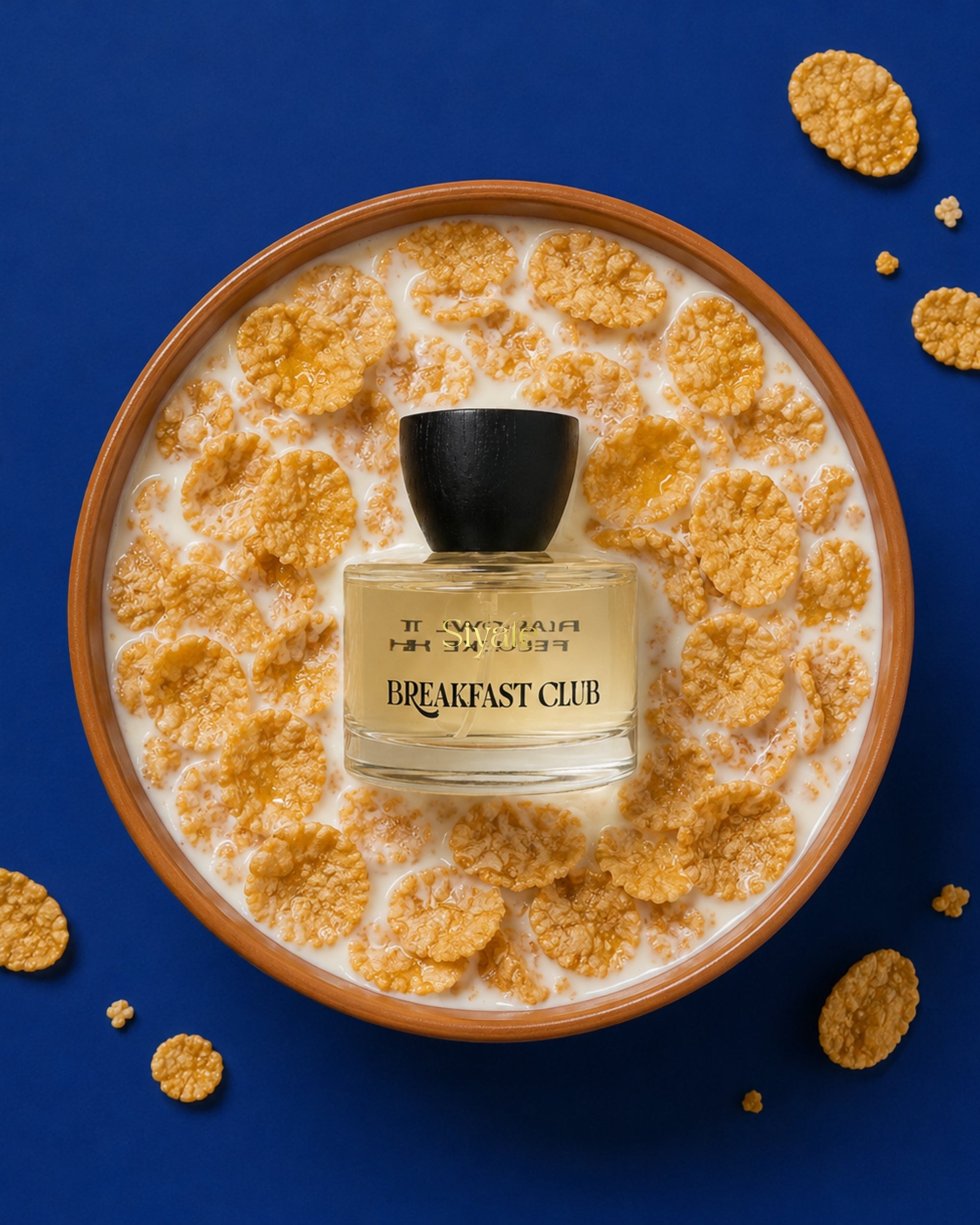 A perfume bottle labeled 'J'ENVOI MAINTENANT BREAKFAST CLUB' sits centered in a bowl of cereal and milk on a deep blue background, with scattered cereal pieces around the edges.