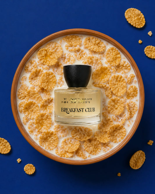 A perfume bottle labeled 'J'ENVOI MAINTENANT BREAKFAST CLUB' sits centered in a bowl of cereal and milk on a deep blue background, with scattered cereal pieces around the edges.