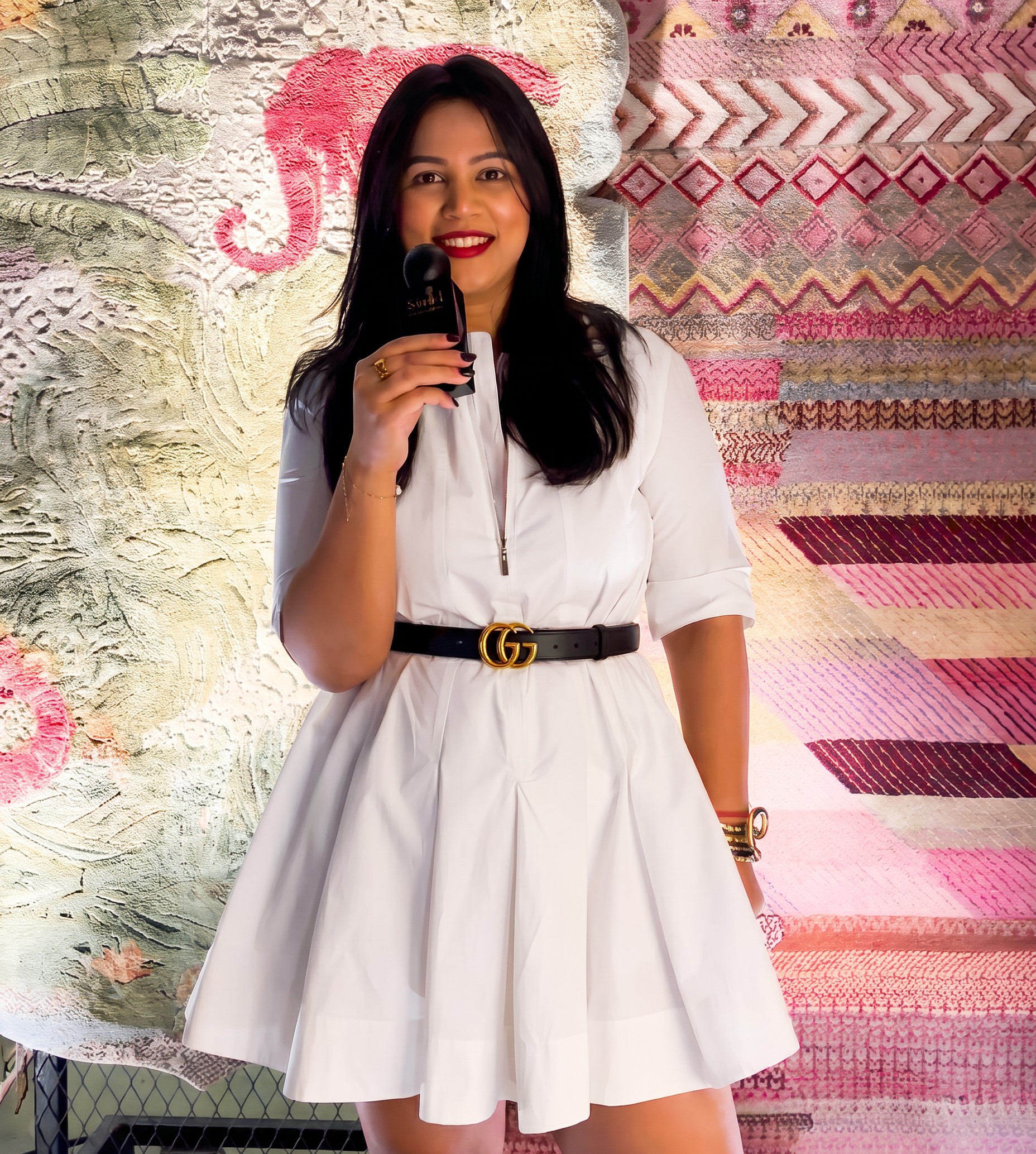A woman wearing a white short-sleeve mini dress with a black belt featuring a gold interlocking G logo, holding a black microphone in front of a patterned textile backdrop.