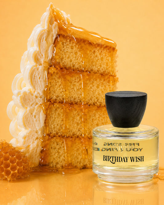 A perfume bottle labeled 'BIRTHDAY WISH' with a black cap, placed in front of a large slice of layered cake dripping with honey, on a warm yellow background.