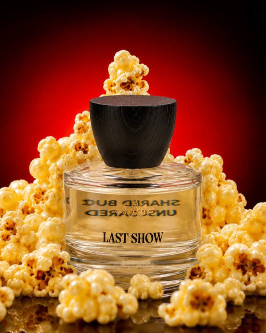 A clear glass perfume bottle labeled 'LAST SHOW' with a black cap, surrounded by popcorn against a dark red gradient background.