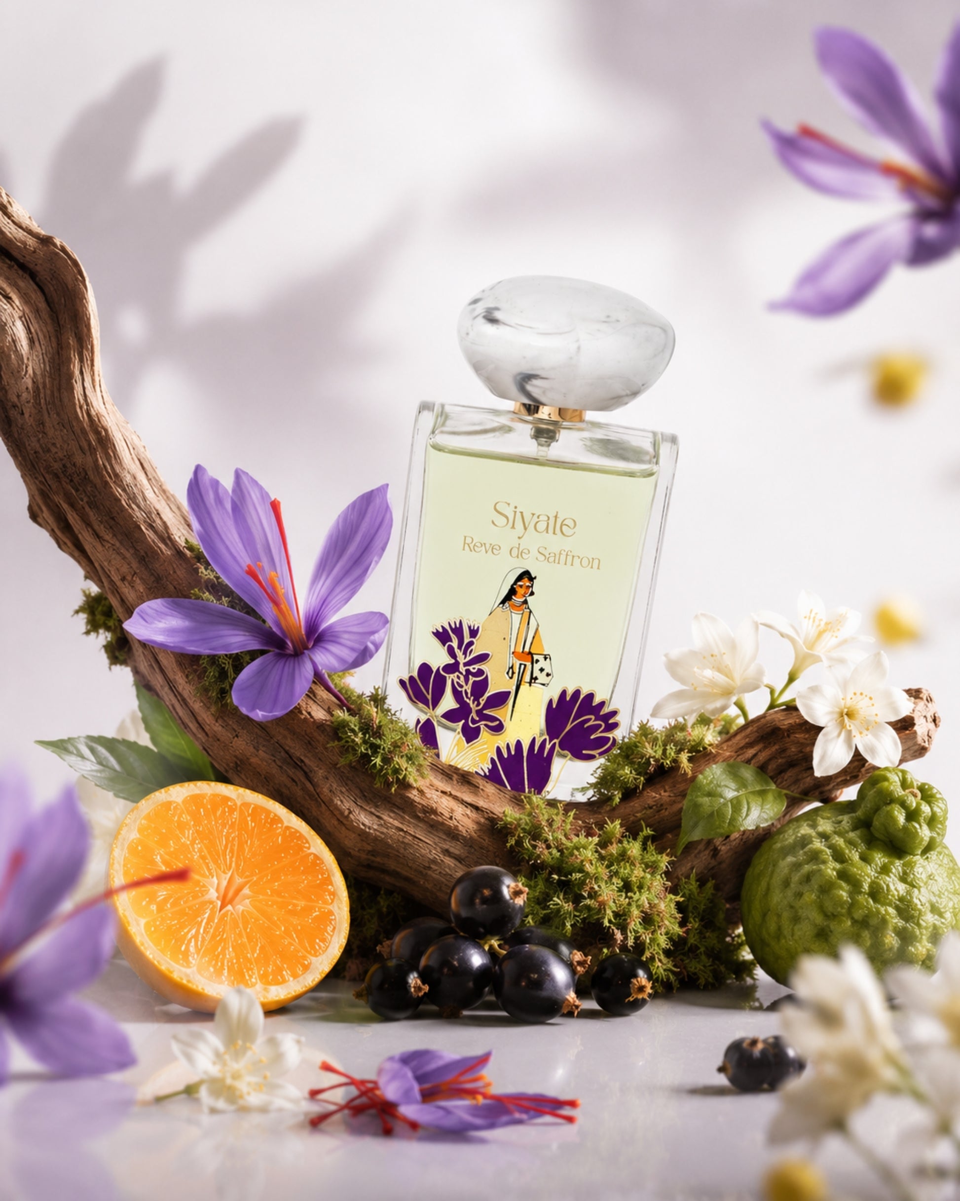 A perfume bottle labeled 'Siyate Reve de Saffron' is styled on a white surface with a curved piece of driftwood, purple flowers, white blossoms, a sliced orange, dark berries, and greenery against a soft white background.