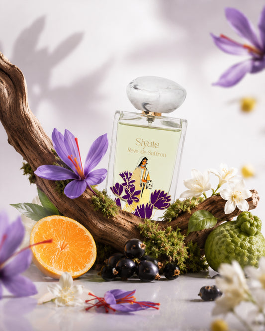 A perfume bottle labeled 'Siyate Reve de Saffron' is styled on a white surface with a curved piece of driftwood, purple flowers, white blossoms, a sliced orange, dark berries, and greenery against a soft white background.