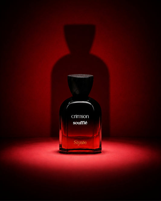 A dark glass perfume bottle labeled 'crimson souplé' and 'Siyate' stands centered on a red-lit surface against a matching red background, with a large shadow behind it.